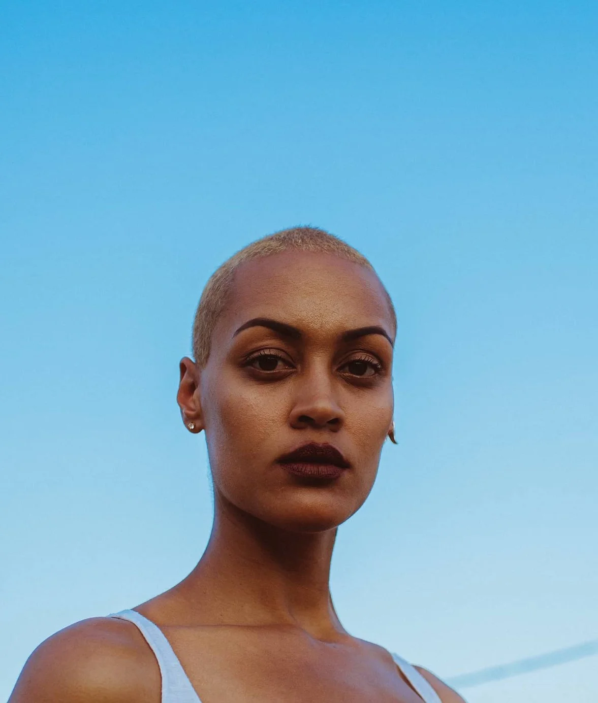 Close-up of a woman with short blonde hair, dark makeup, wearing earrings, against a clear blue sky.