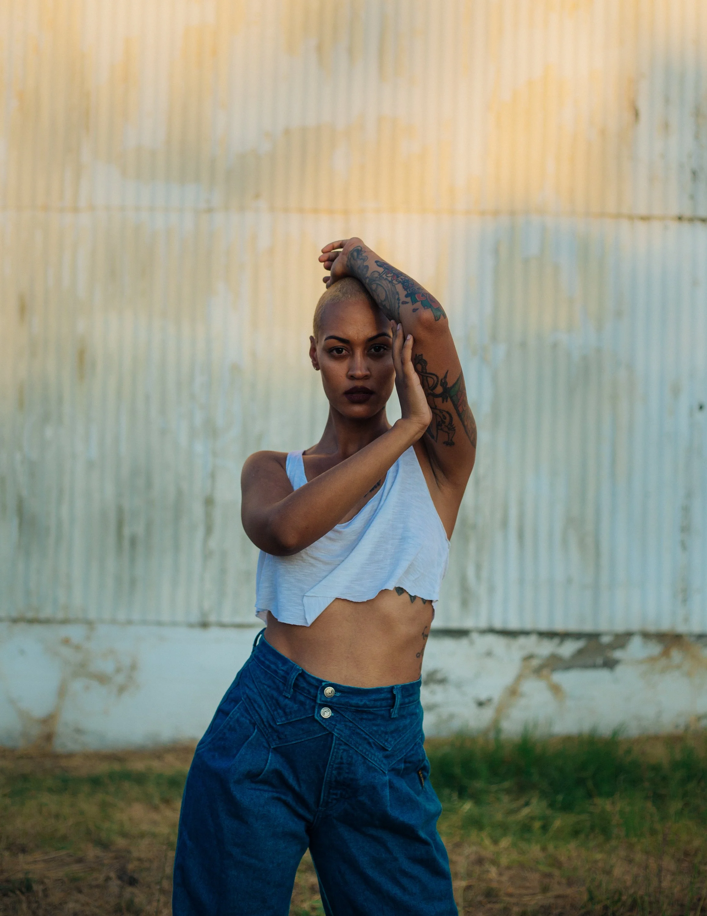 A woman with tattoos and a shaved head posing outdoors against a corrugated metal wall, wearing a white crop top and high-waisted blue jeans.