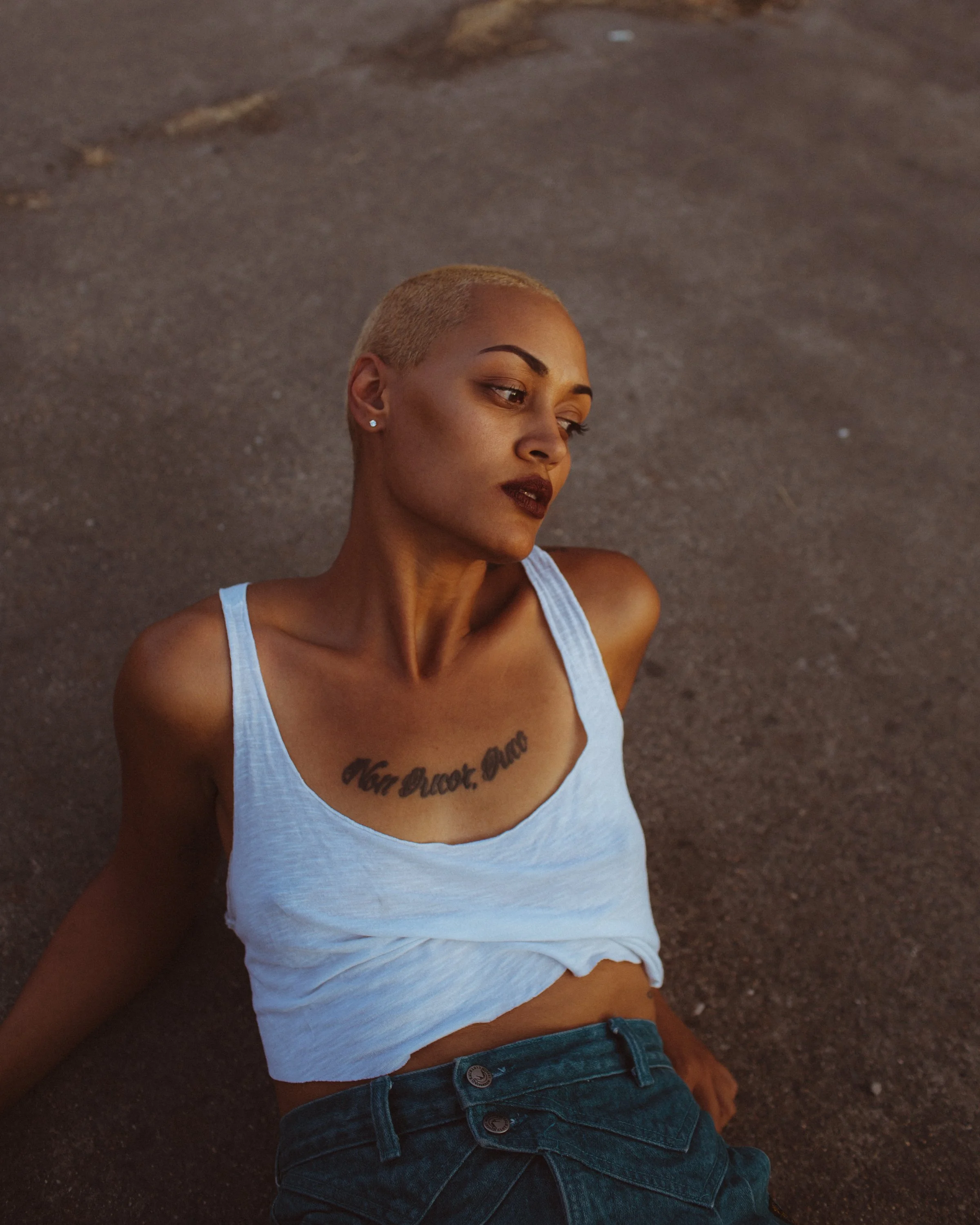 A woman with short blonde hair and dark lipstick sitting on the ground with a white tank top and dark jeans.