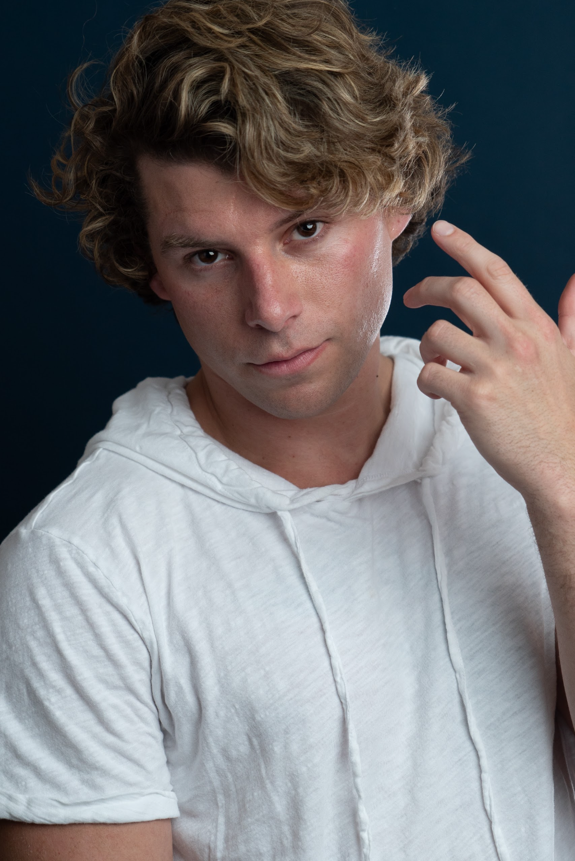 Travis Grant with curly blonde hair wearing a white hoodie, looking directly at the camera with a slight smirk, against a dark background.