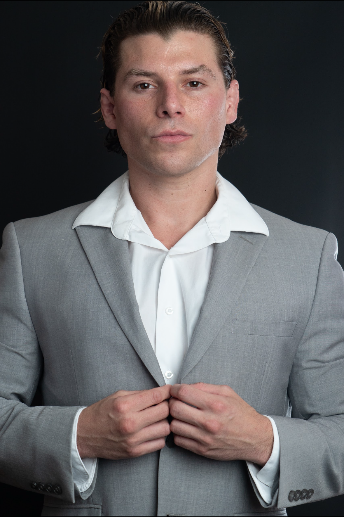 Travis Grant with medium-length dark hair, wearing a light grey suit and white dress shirt, is standing against a dark background, looking directly at the camera with a serious expression, adjusting his suit jacket.