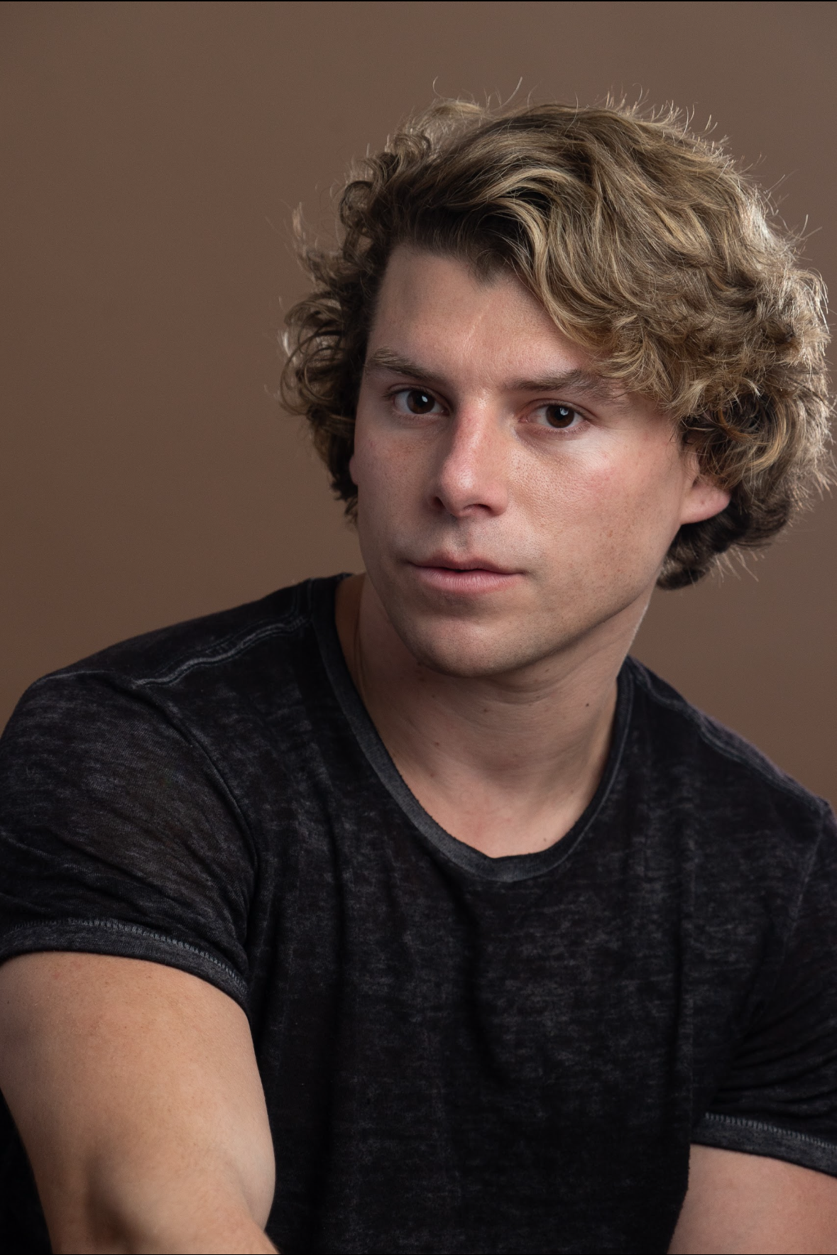 Travis Grant with curly blonde hair and fair skin wearing a black T-shirt, posing against a brown background.