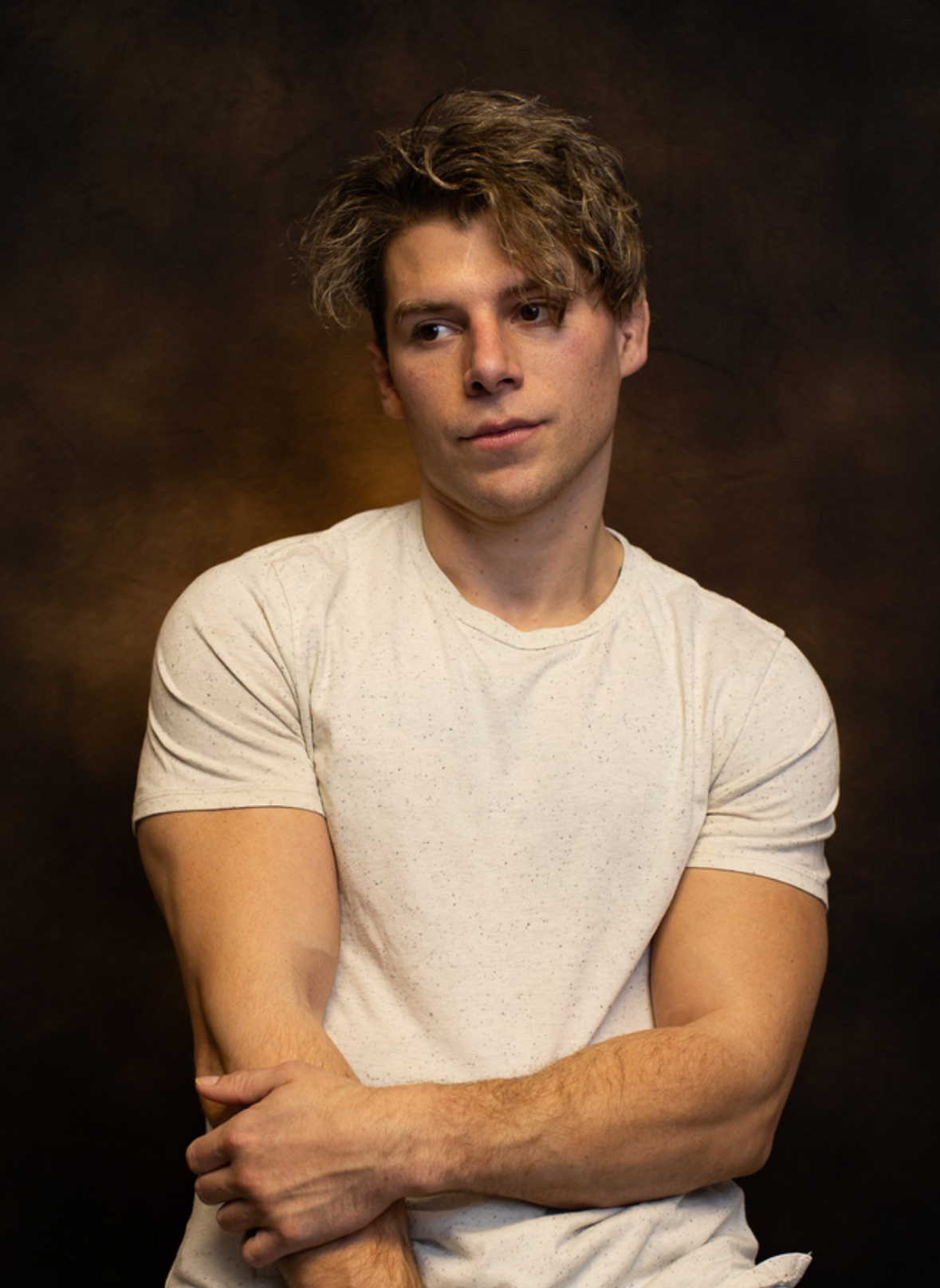 Portrait of Travis Grant with messy, wavy hair, wearing a white speckled T-shirt, against a dark brown background.