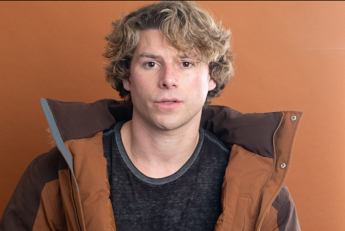 Travis Grant with curly blond hair, wearing a black shirt and a brown jacket, standing against a brown background.