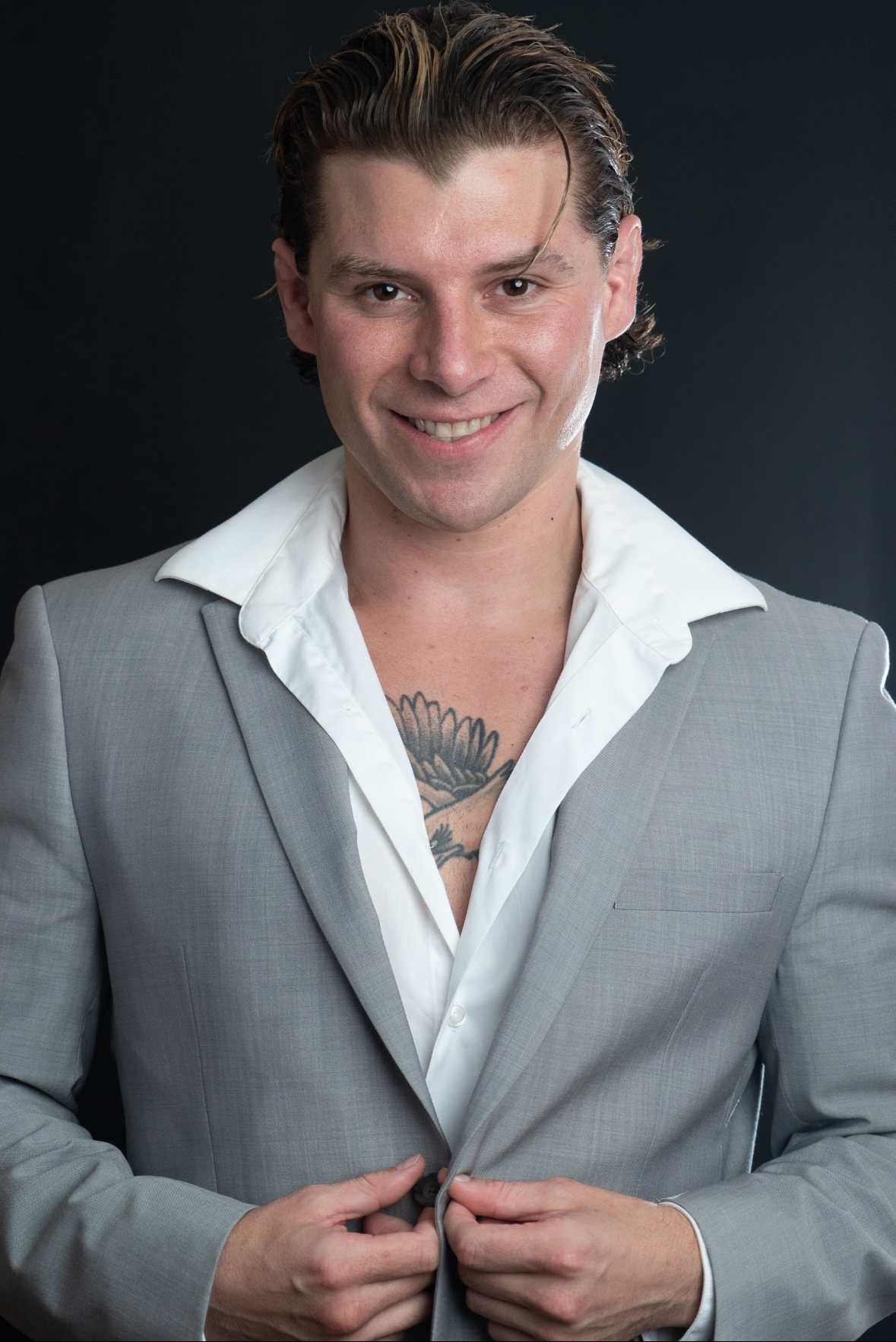 Travis Grant with dark, slicked-back hair, smiling and wearing a light gray suit jacket over a white dress shirt, with a tattoo visible on his chest, against a dark background.