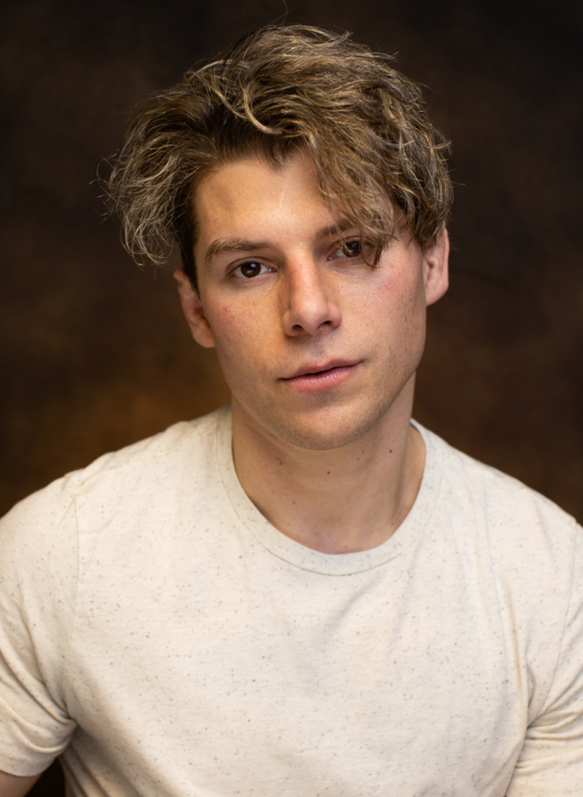 A young man with wavy, tousled hair and a slight smile, wearing a light-colored, speckled t-shirt, standing against a dark, textured background.