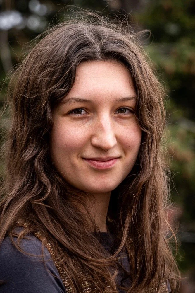 Close-up of a young woman with long, wavy brown hair and a subtle smile, outdoors with blurred greenery in the background.