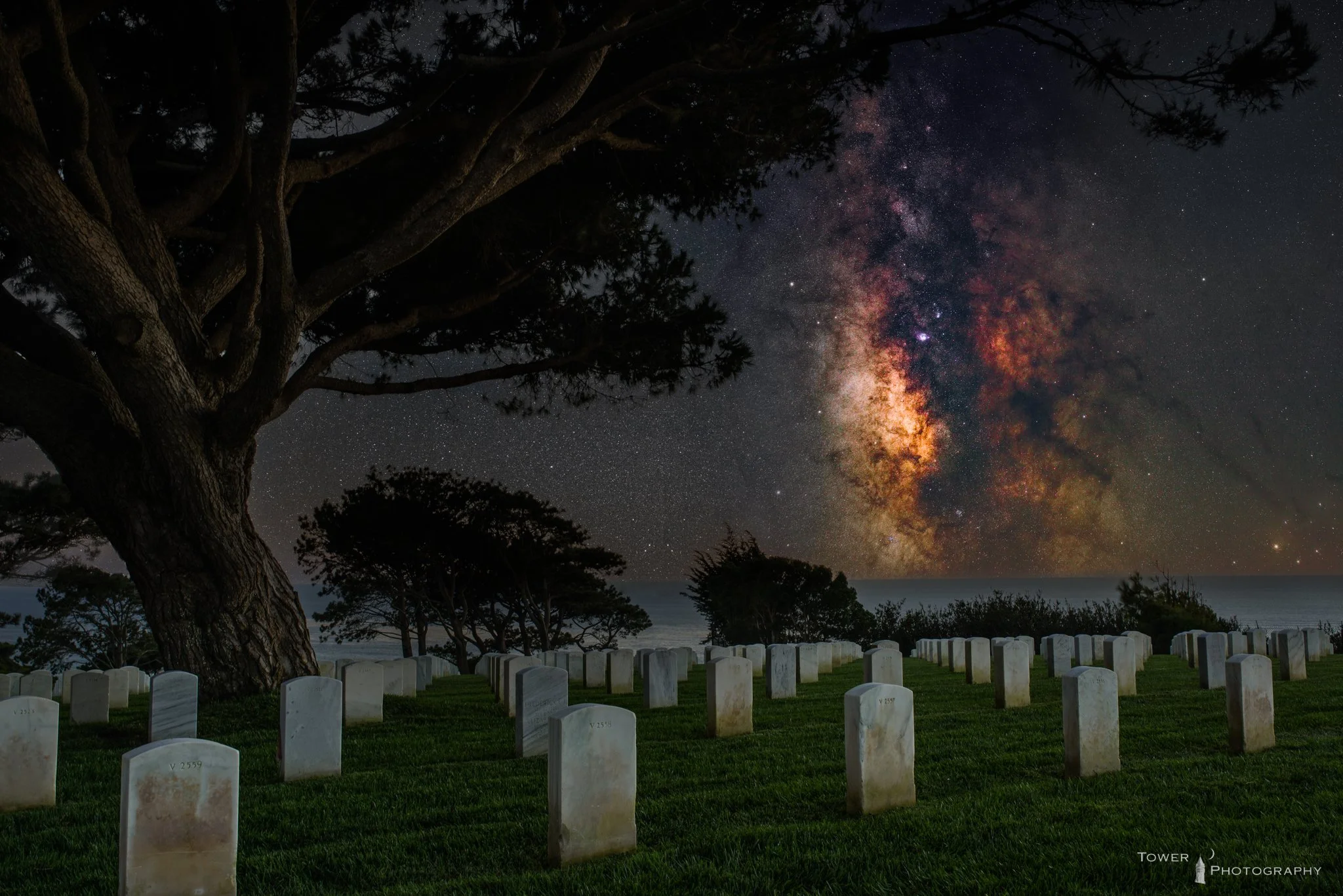 Nighttime scene of a cemetery with rows of white gravestones beneath a large tree. The sky is clear with a view of the Milky Way galaxy, showing bright stars and colorful cosmic dust.