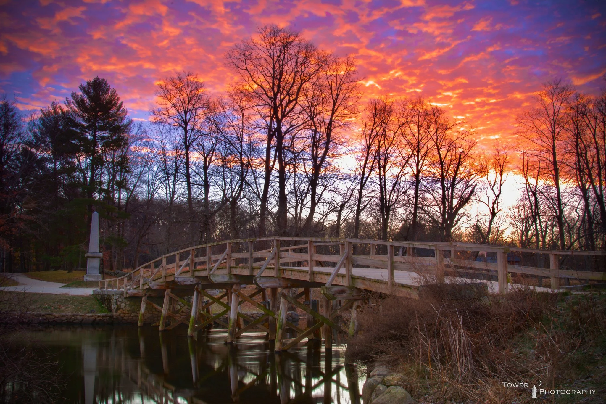 Wooden bridge over a small river with a sunset sky filled with orange and purple clouds and bare trees in the background.