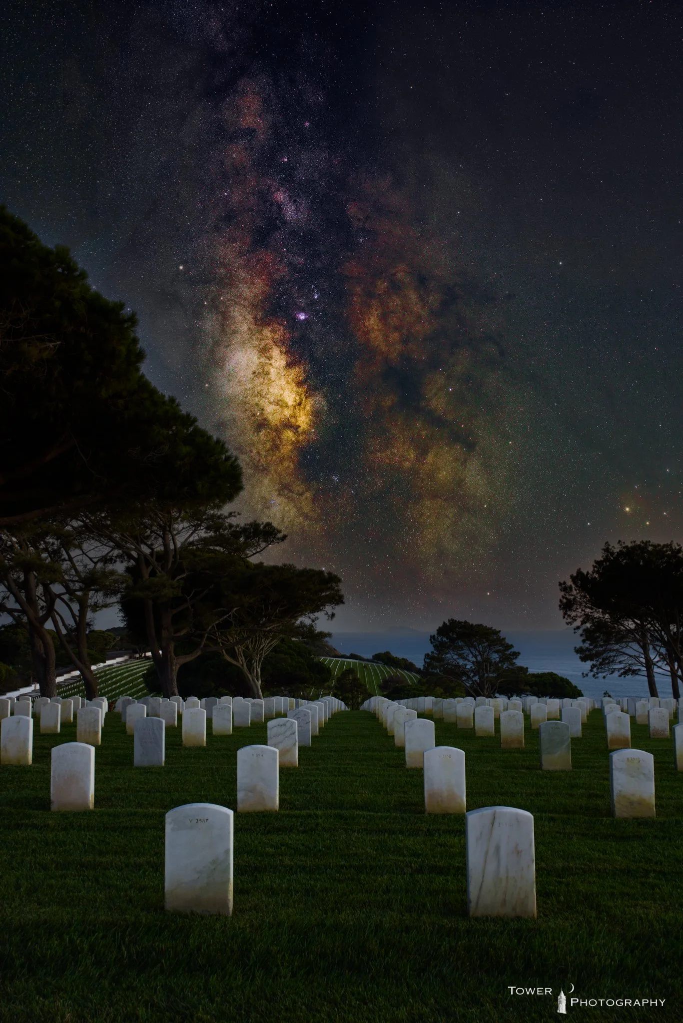 Nighttime scene of a cemetery with rows of white headstones, surrounded by trees, under a clear night sky filled with stars and the Milky Way galaxy.