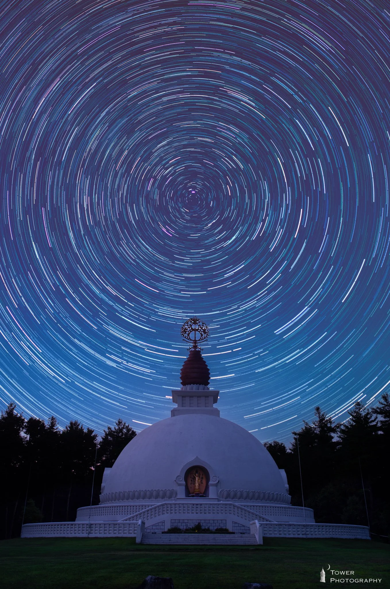 Long exposure photo of star trails swirling above a white Buddhist stupa with a small statue inside, surrounded by trees at night.