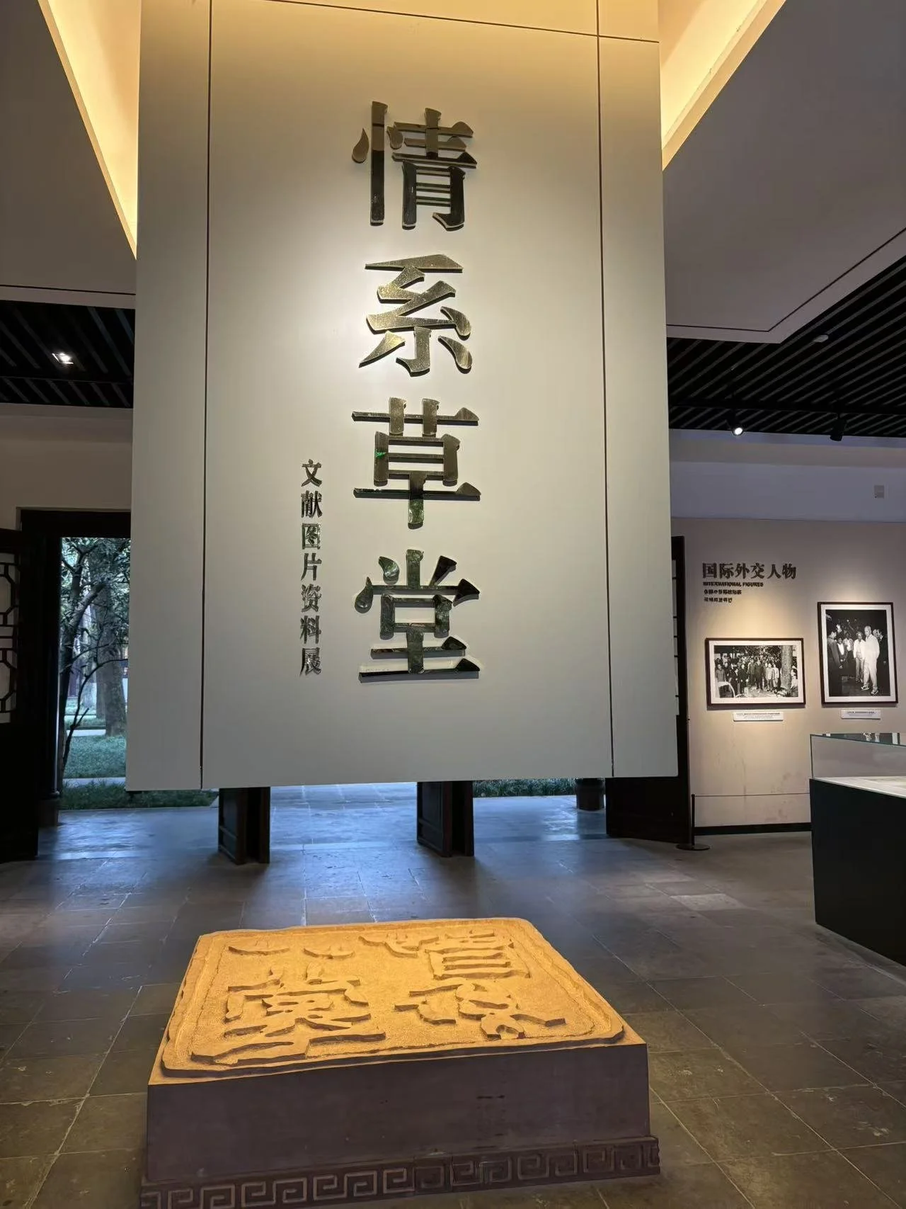 Large vertical sign with Chinese characters on a white wall, and a carved wooden plaque on a stand in front of it. The sign appears to be in a museum or exhibition space.