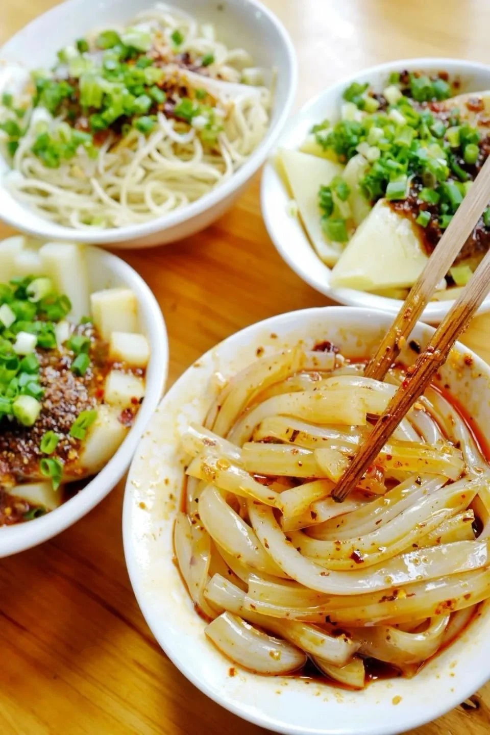 Four bowls of Asian noodle dishes with green onions, sliced vegetables, and sauces on a wooden table.