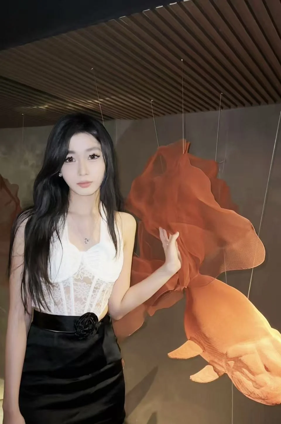 A woman with long black hair and fair skin standing indoors next to a large orange fish sculpture suspended from the ceiling.