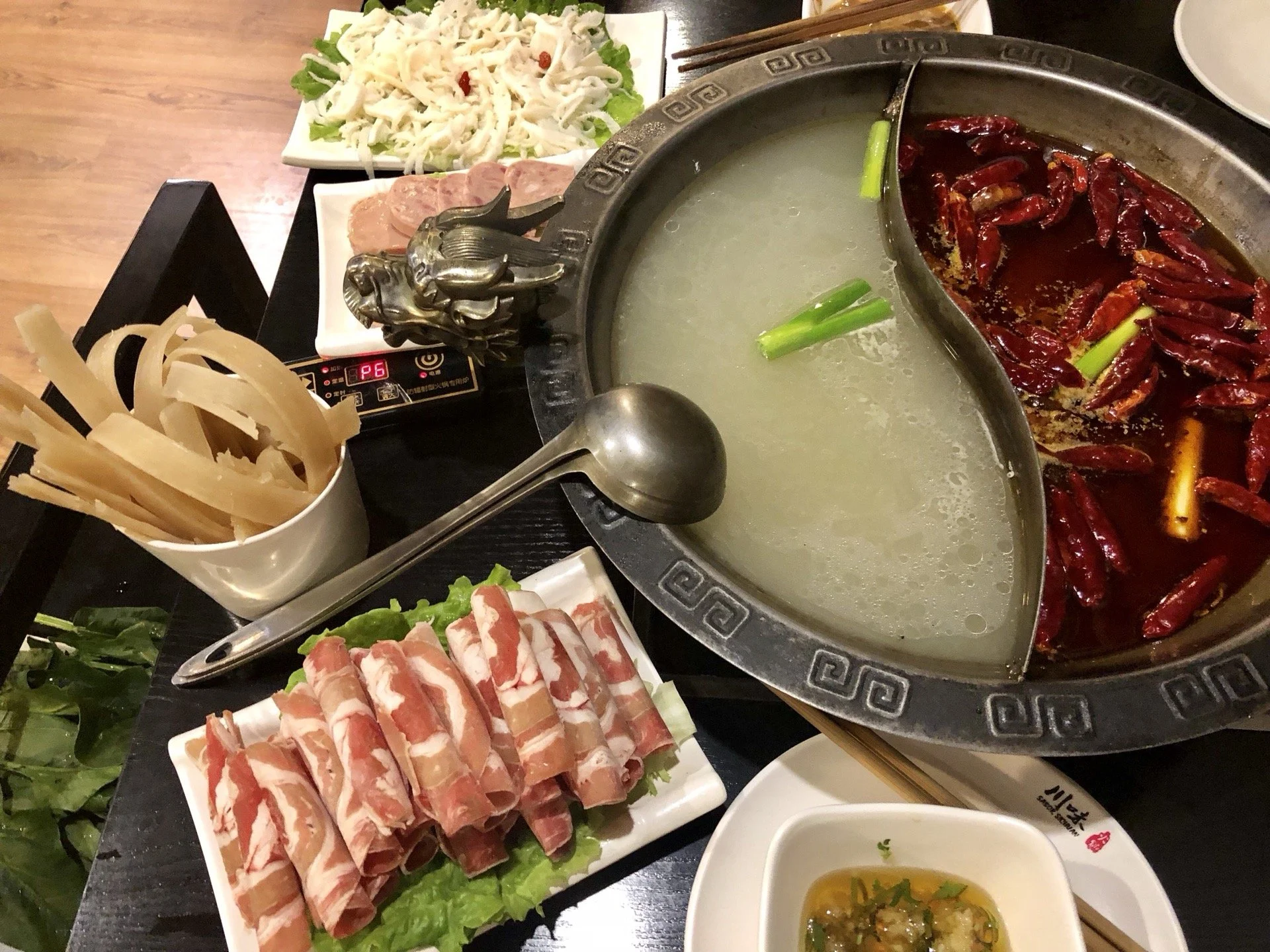 A Chinese hot pot meal with a divided pot containing broth and spicy chili oil, surrounded by plates of sliced meat, vegetables, tofu, and condiments on a black table.