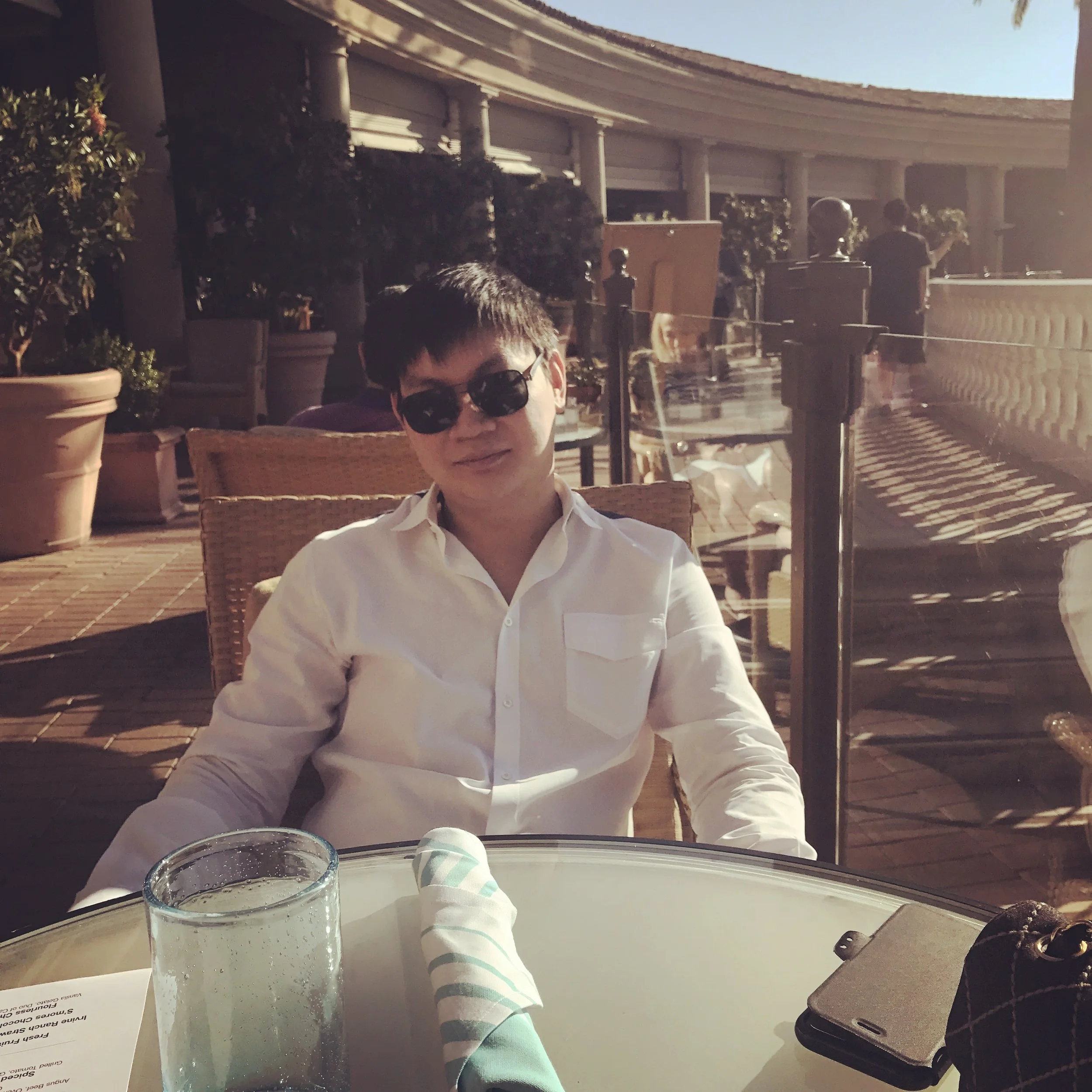 A person wearing sunglasses and a white shirt sitting at an outdoor restaurant table during daylight, with a glass of water, a striped napkin, a phone, and a purse on the table, surrounded by potted plants and other diners.