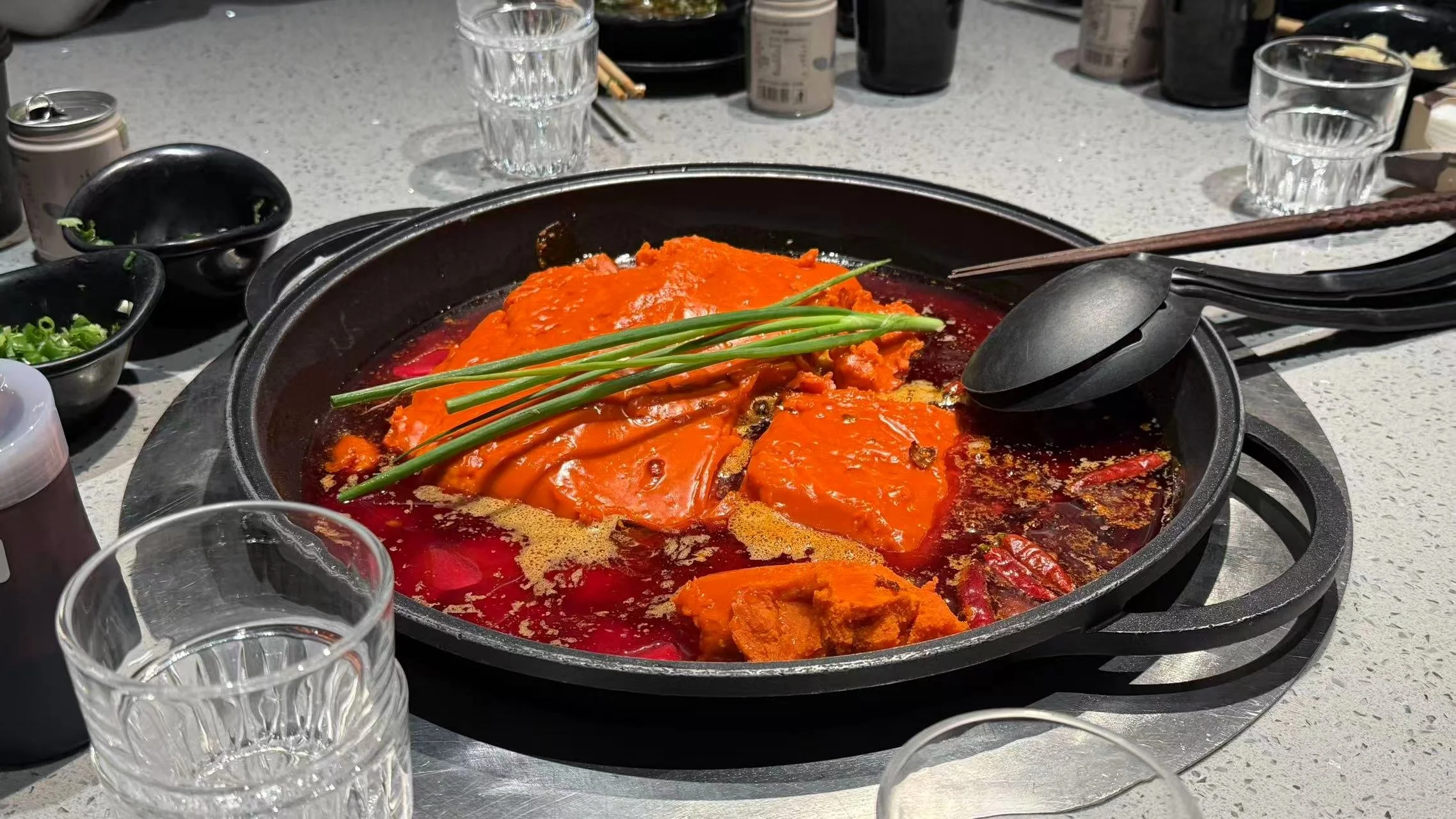 A sizzling hot pot with red broth and chunks of meat, garnished with green onions, surrounded by glasses and bowls on a gray tabletop.