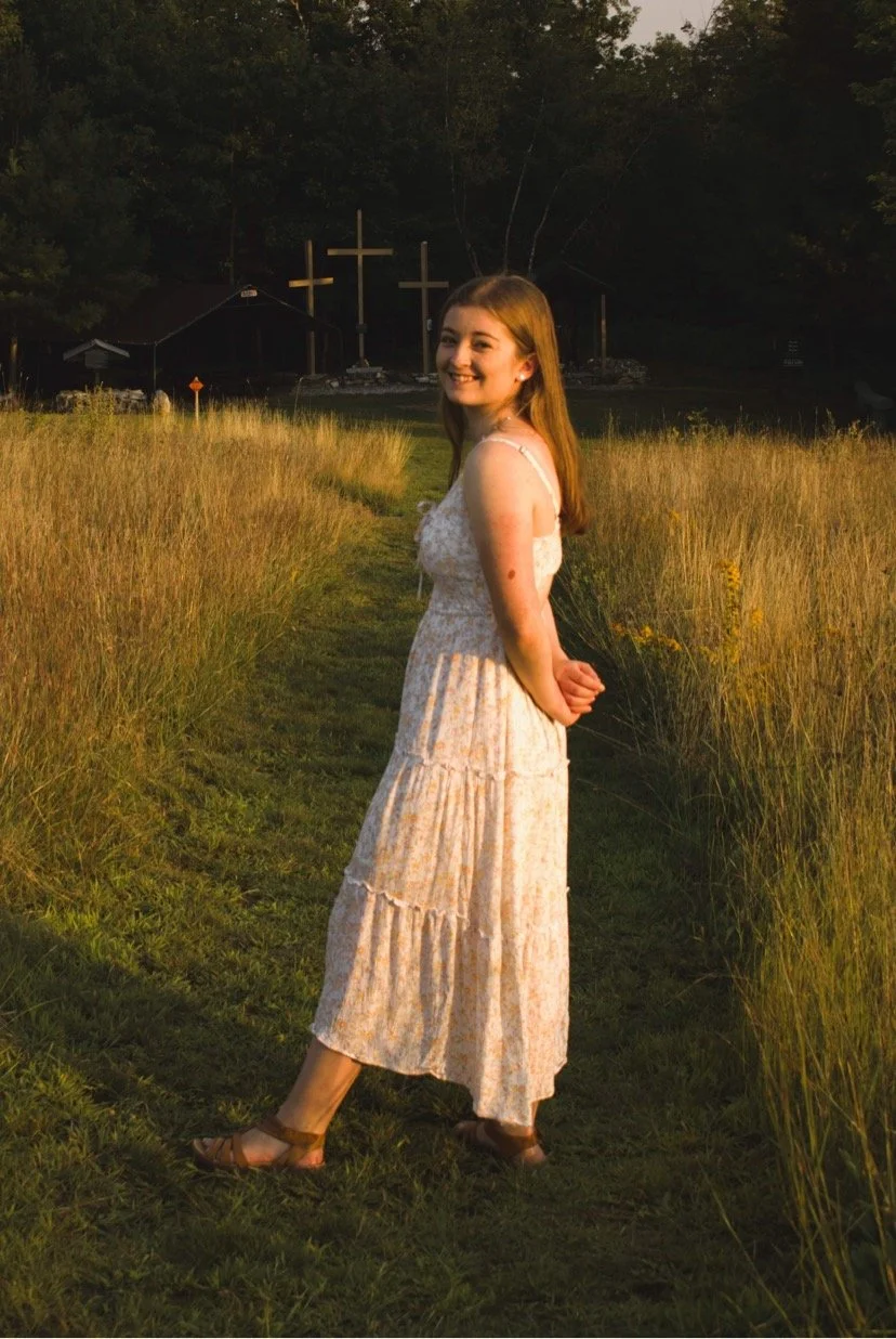 A young woman with light skin and long brown hair standing on a grassy path in a field, smiling at the camera. Behind her, there are three crosses in front of a small building, with trees in the background.