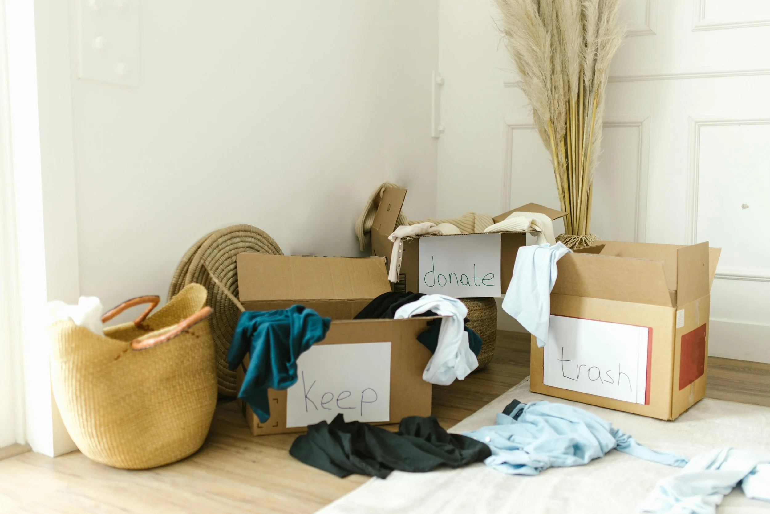 Several open cardboard boxes with laundry and clothes spilling out, labeled 'donate,' 'keep,' and 'trash,' placed on a wooden floor near a white wall and a bunch of tall dried pampas grass.