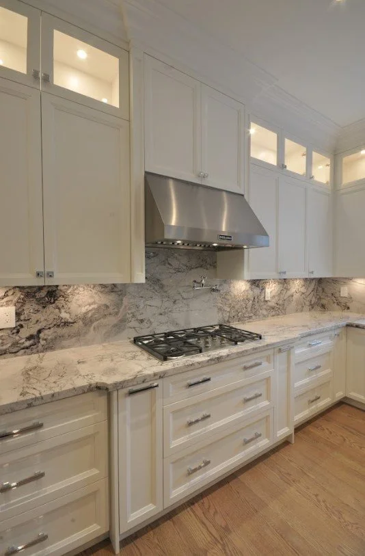 White kitchen with marble countertops, upper glass-front cabinets, and a stainless steel range hood over a gas cooktop.