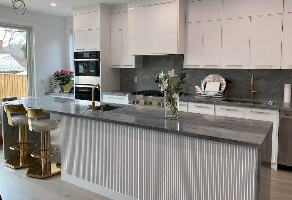 Modern kitchen with white cabinetry, gray marble countertops, and gold hardware. A kitchen island features a gray marble top and gold bar stools. There is a built-in oven, a gas cooktop, a gold faucet, and pink flowers on the island. A window provide