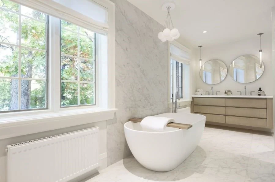Modern bathroom with freestanding oval bathtub, marble walls and floors, large windows with blinds, double vanity with round mirrors, and hanging pendant lights.