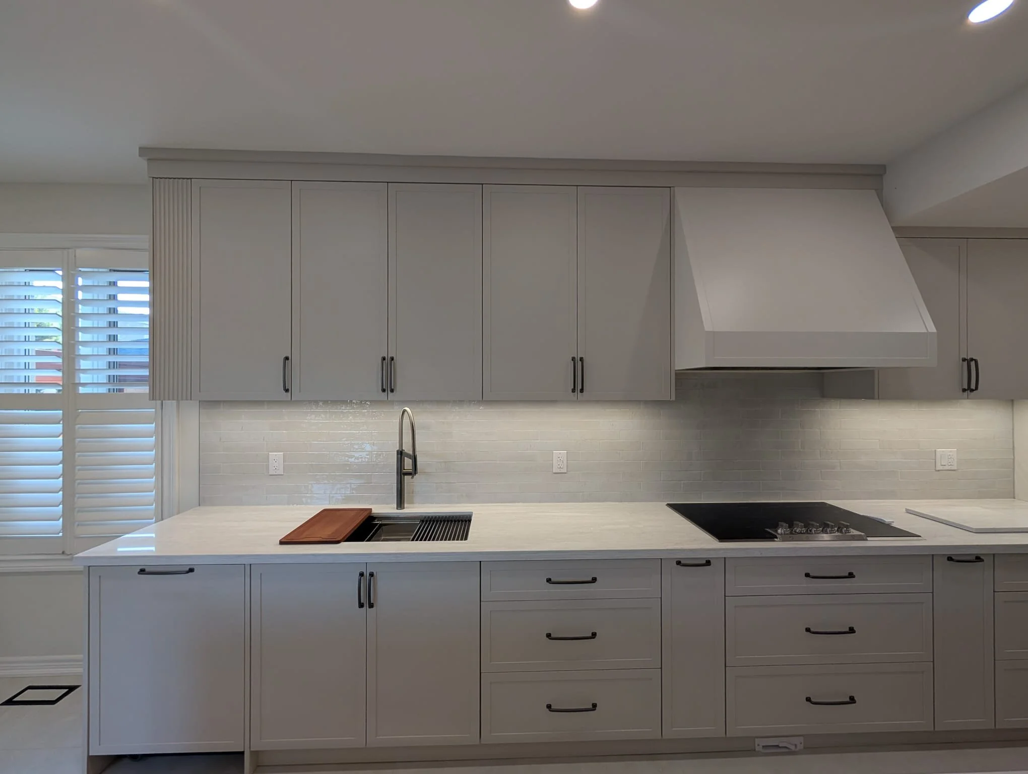Modern kitchen with white cabinets, a white countertop with a wooden cutting board, a stainless steel sink with a faucet, a black cooktop, and a window with white shutters.