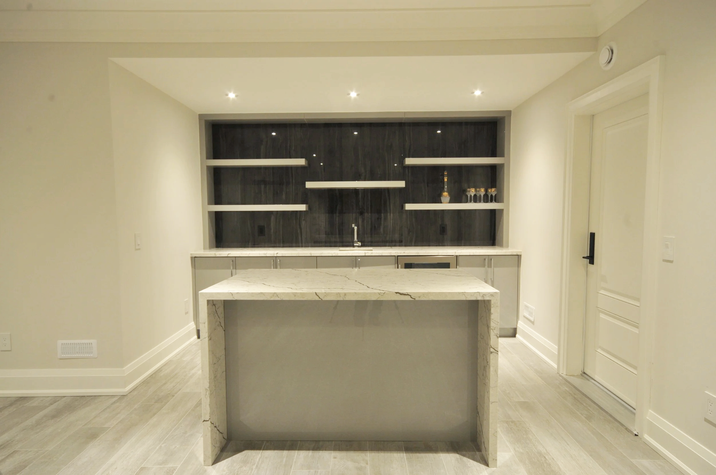 Modern kitchen with white marble island, dark wood backsplash, and white floating shelves, with a small bar area and glassware.