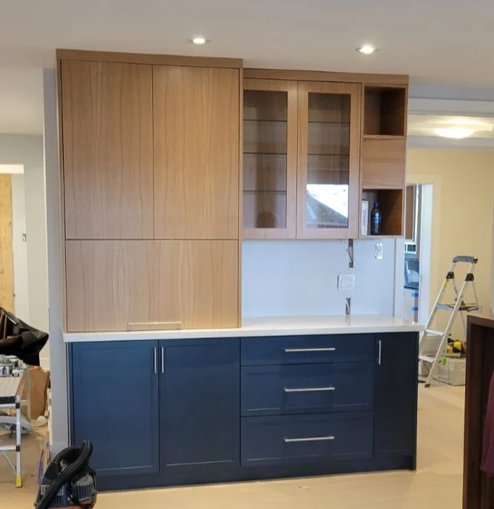 Partially installed kitchen cabinets with a combination of natural wood upper cabinets, some with glass doors, and dark blue lower cabinets with silver handles, set against a light-colored backsplash and countertop.