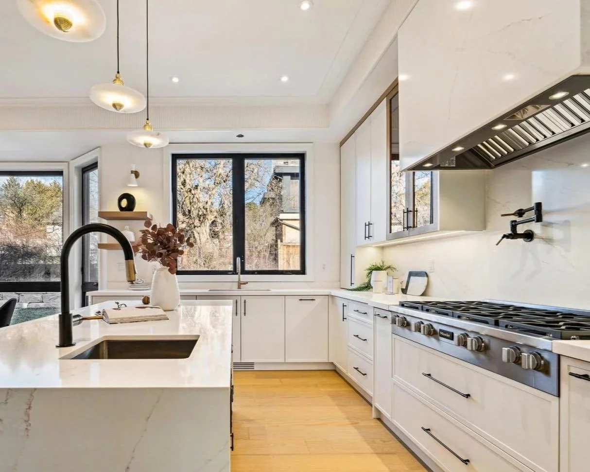 Modern kitchen with white cabinets, a large marble island, black faucet, stainless steel stove, and large windows showing trees outside.