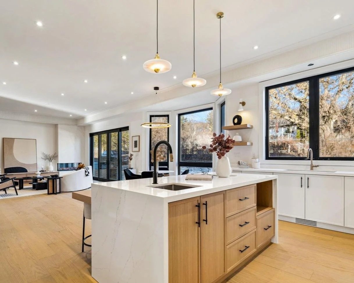 Modern open-concept kitchen and living room with large windows, wooden flooring, white cabinetry, and a central island with a black faucet, decorated with a vase of dried flowers.