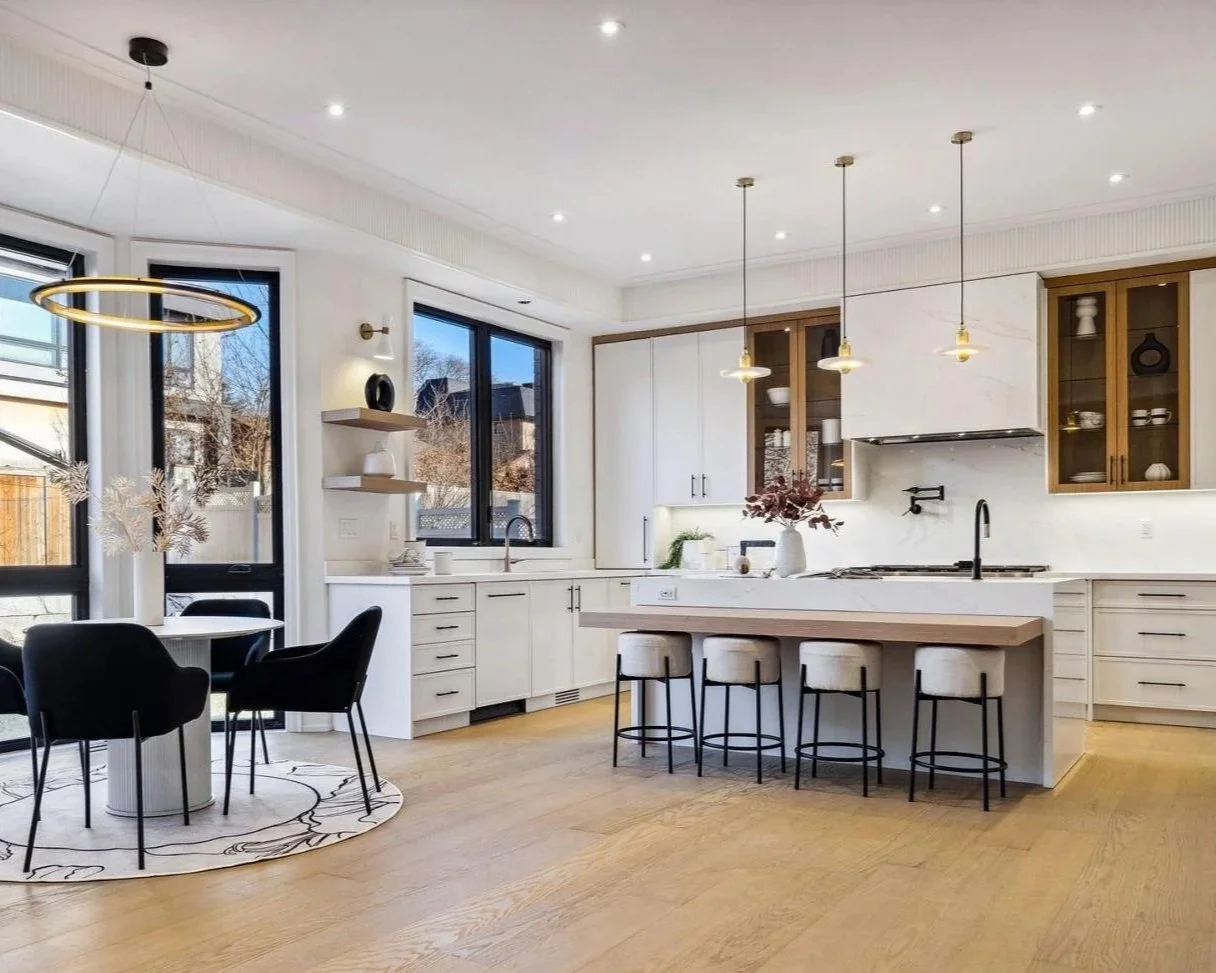 Modern kitchen with white cabinets, wooden accents, a kitchen island with four barstools, black fixtures, and a round dining table with black chairs near large windows.