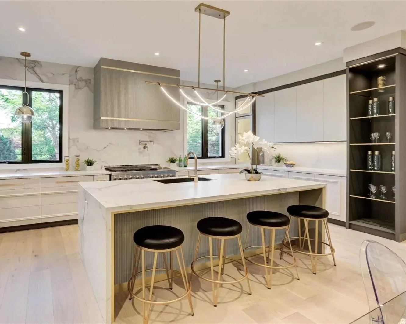 Modern kitchen with white marble island, black bar stools, gray cabinets, and open shelving display glasses, with large windows showing greenery outside.
