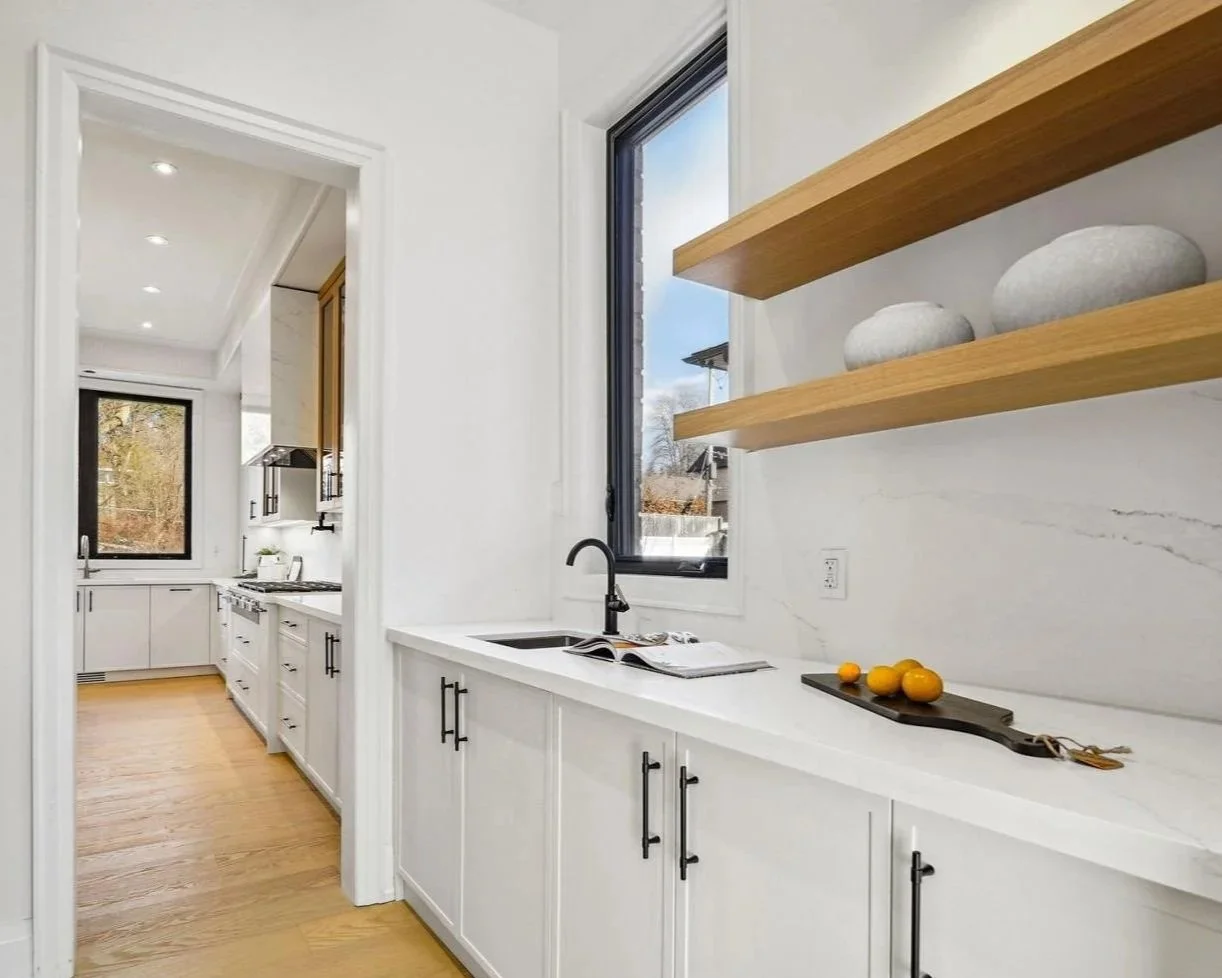 Modern kitchen with white cabinets, black hardware, wooden open shelves, black window frames, and a black faucet, with natural light coming through large windows.
