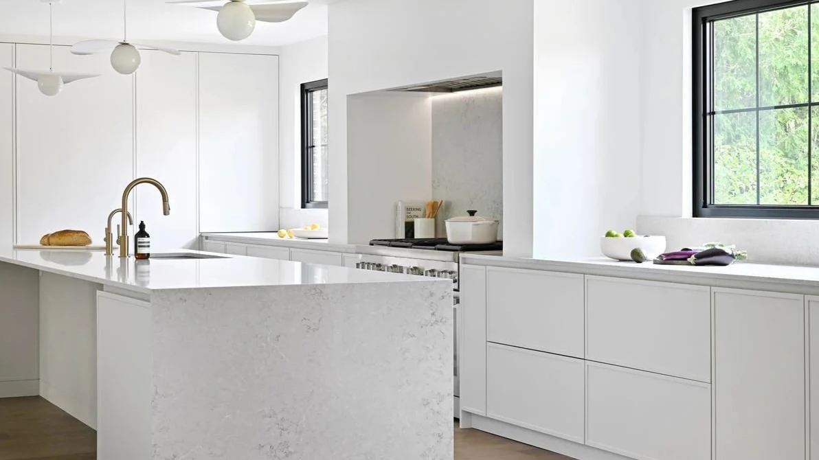 Modern white kitchen with black-framed windows, white countertops, and built-in appliances, decorated with bowls of fruit and kitchen utensils.