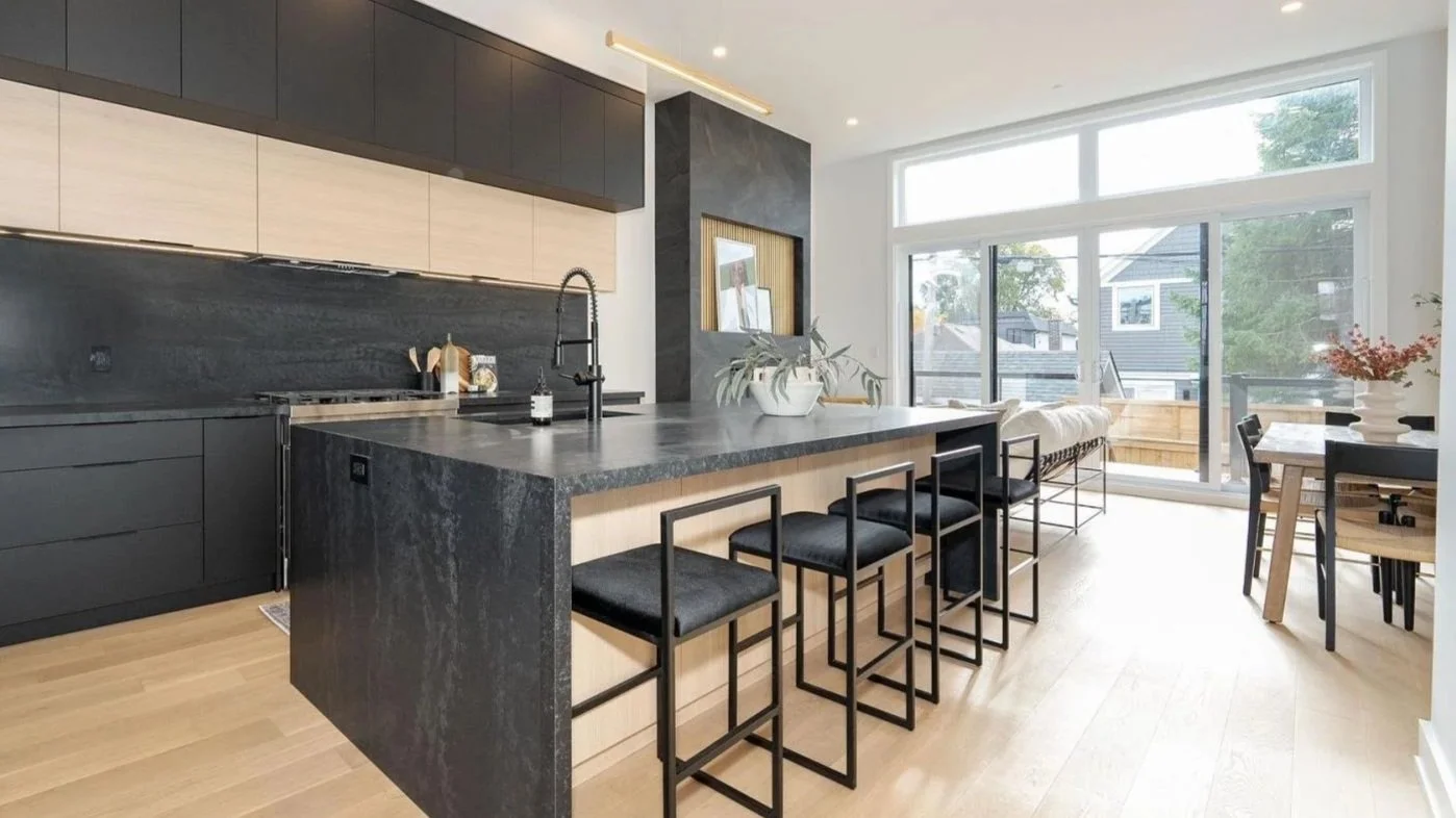 Modern kitchen and dining area with large windows and natural light, featuring black and wood cabinets, a black stone island with seating, and a dining table with chairs, decorated with potted plants and a vase of flowers.