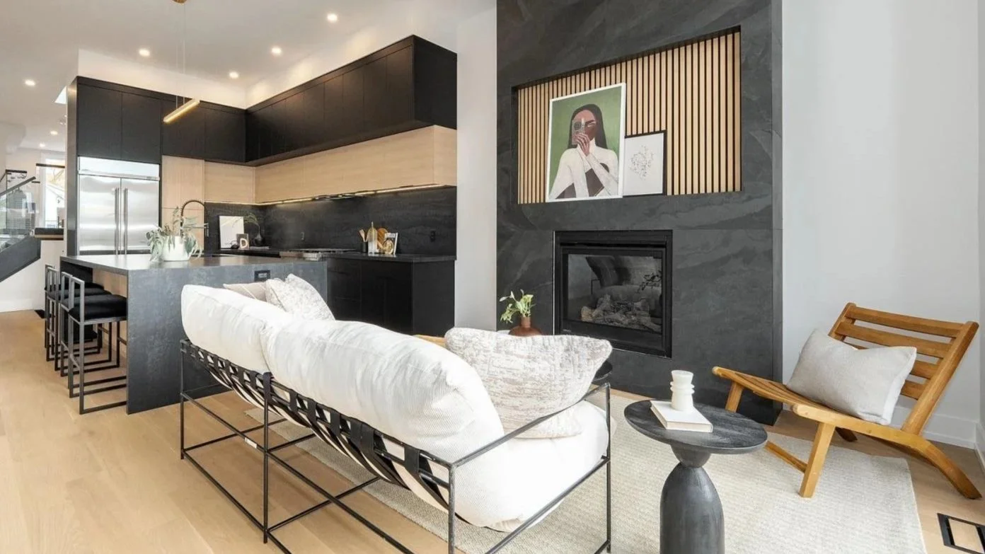 Modern open-concept living room with a black and beige kitchen in the background. The seating area includes a white sofa with pillows, a wooden lounge chair with a pillow, and a small black round side table with a book and cup. A sleek black fireplac