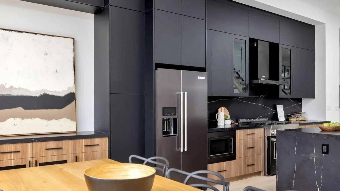 Modern kitchen with black cabinets, stainless steel refrigerator, wooden cabinets, black marble countertops, and some decorative items.