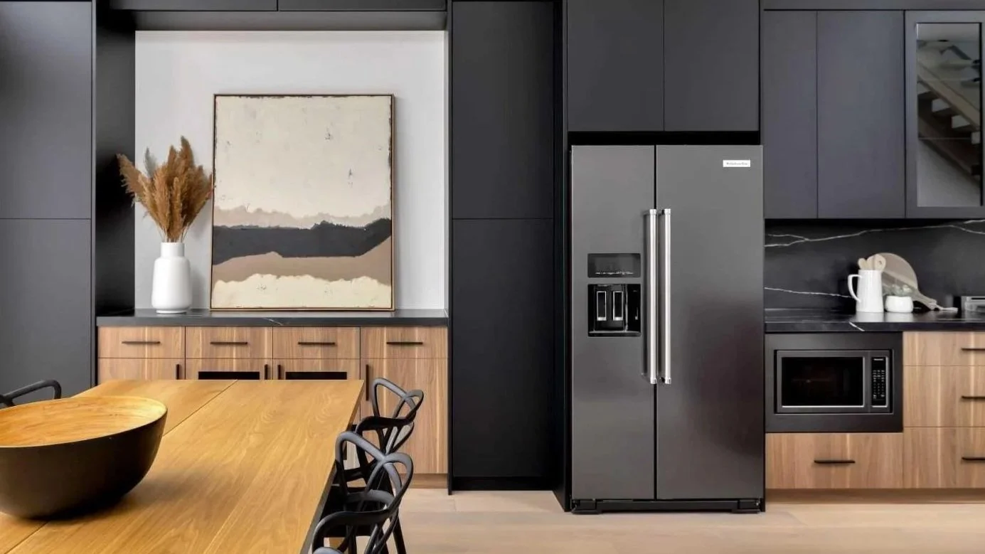 Modern kitchen with black cabinets, stainless steel refrigerator, wooden drawers, a black bowl on a wooden dining table, and decorative artwork with a vase and dried plants.