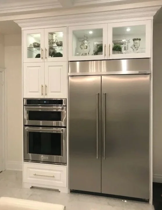 White kitchen cabinetry with glass front upper cabinets, stainless steel double ovens, and a side-by-side stainless steel refrigerator.