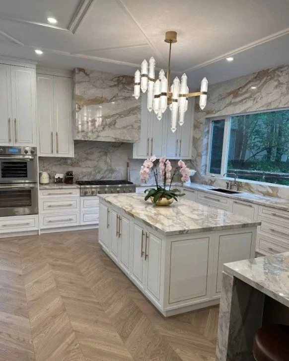 Modern kitchen with white cabinetry, marble backsplash, and island with marble countertop, featuring a hanging chandelier and a large window with green trees outside, and a potted pink orchid on the island.