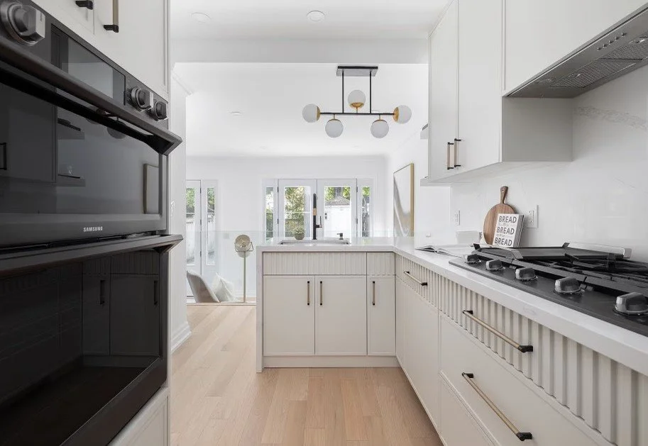 Modern white kitchen with built-in oven, gas stove, white cabinets, and a large window with a view of a patio.