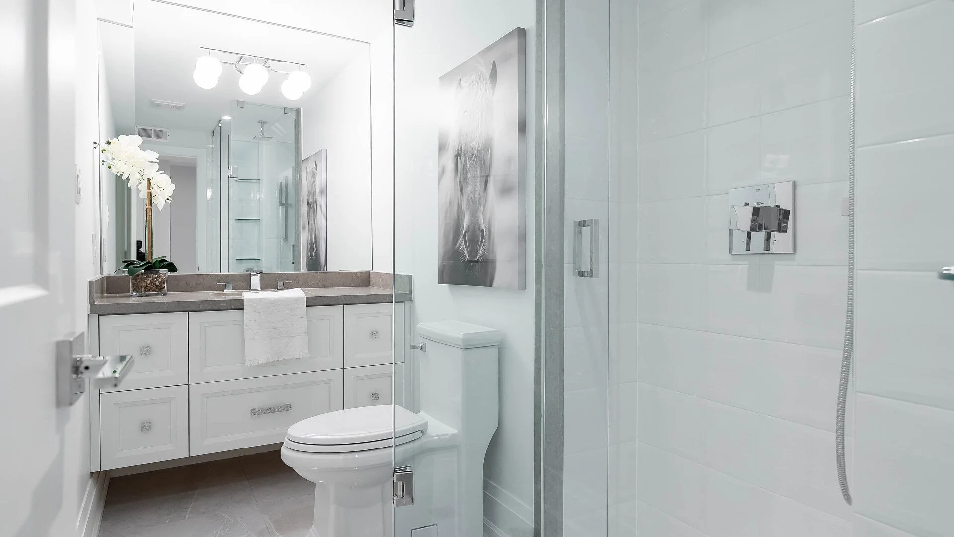 Modern white bathroom with vanity, toilet, and walk-in shower, decorated with a flower in a pot and a horse painting