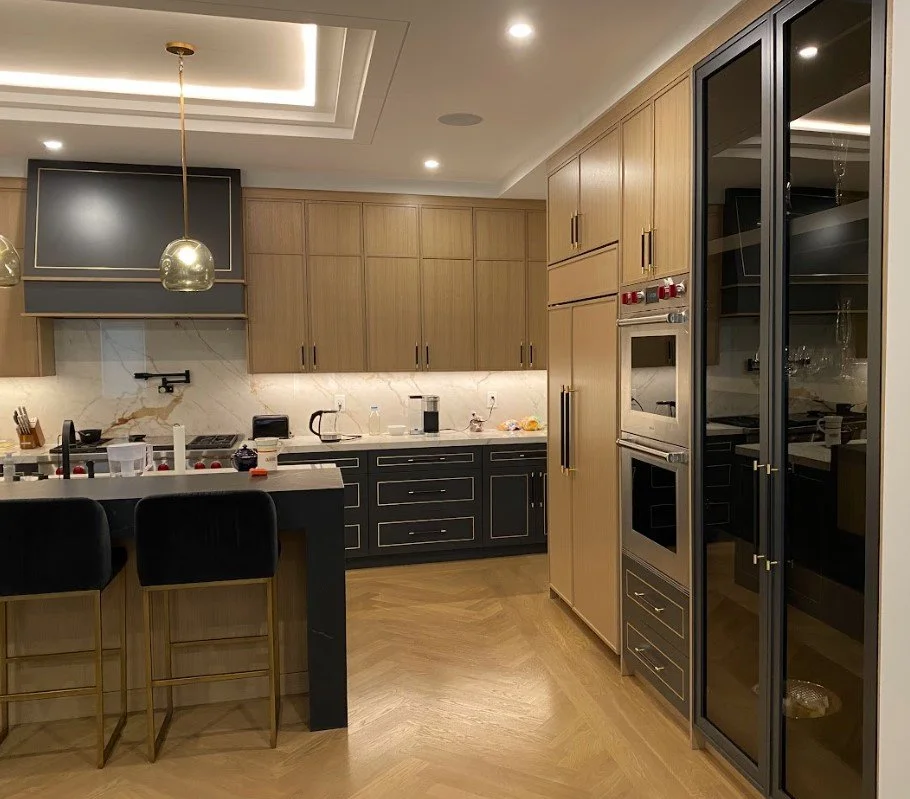 Modern kitchen with wooden cabinets, black lower cabinets with gold handles, built-in double ovens, marble backsplash, black countertops, black bar stools, and a large black glass-fronted pantry or cabinet.