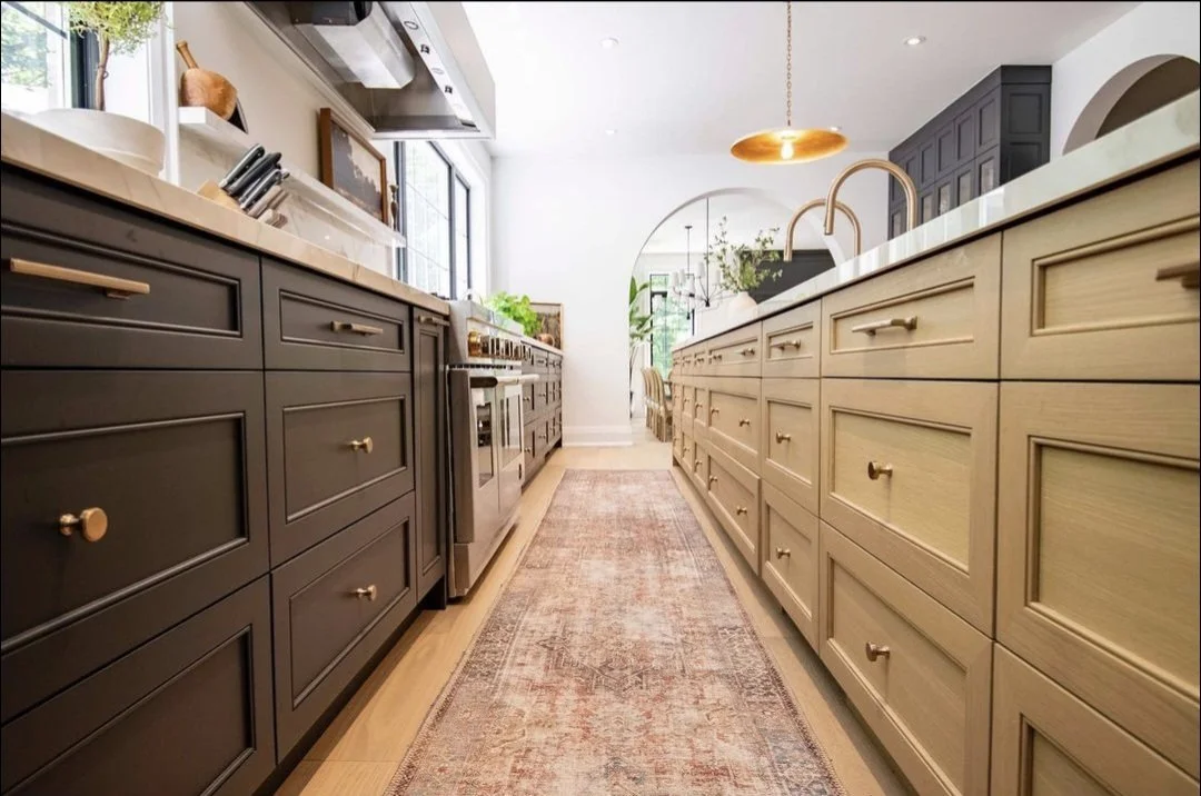 A modern kitchen with a central walkway, dark and light wooden cabinets, a marble countertop, stainless steel appliances, a hanging copper light fixture, and natural light from windows.