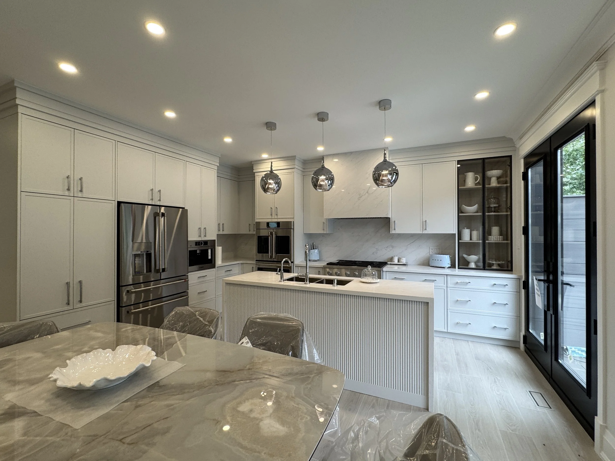 Modern kitchen with white cabinets, stainless steel appliances, marble backsplash, pendant lights, and large sliding glass door leading outside.