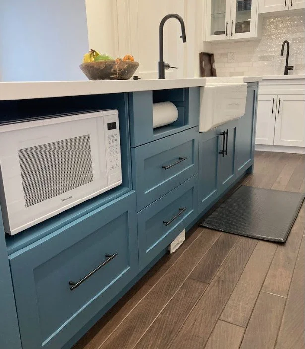 Kitchen with blue cabinets, a white microwave, a black faucet, a black dish towel, and a bowl of fruit on the counter.