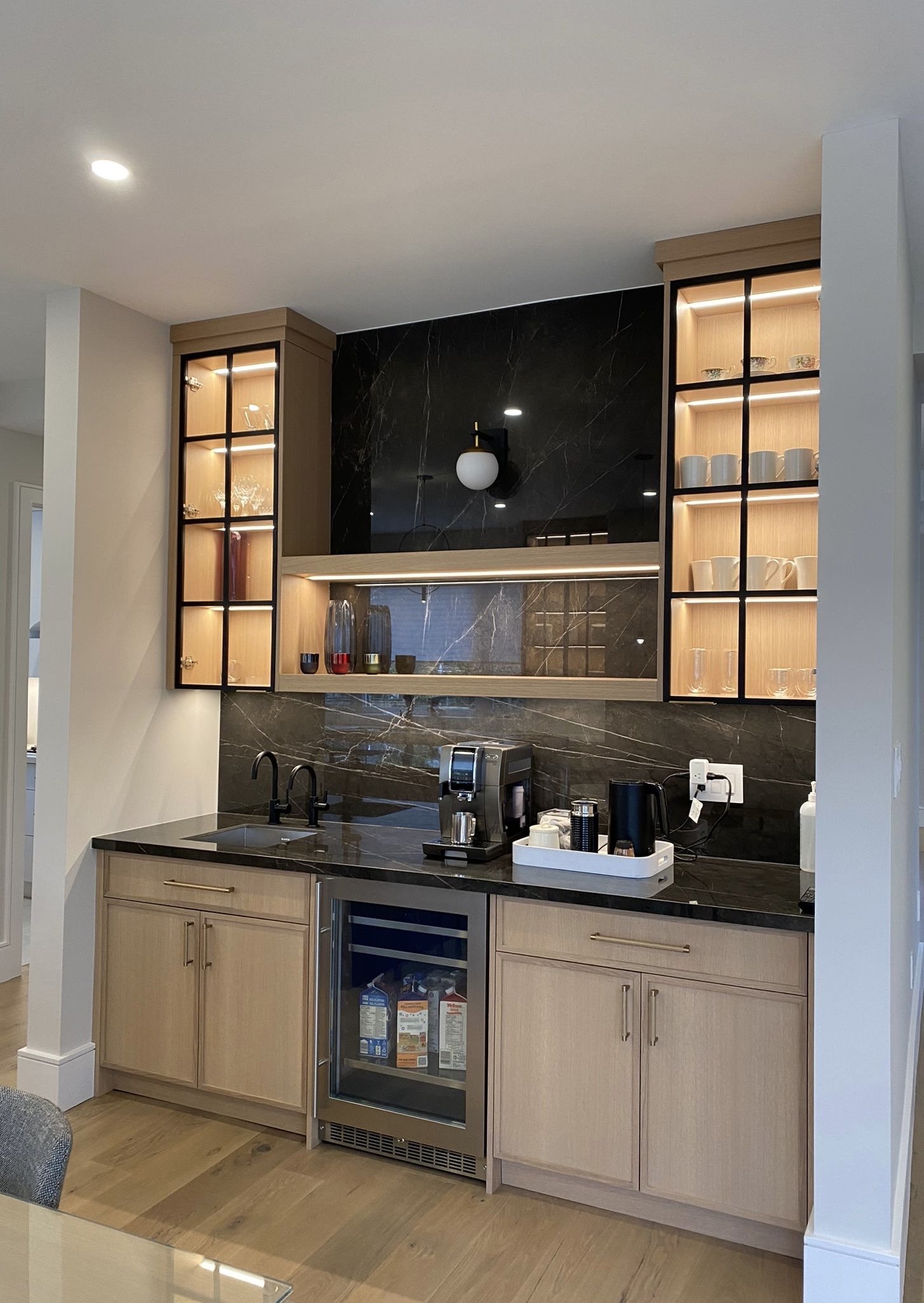 Modern kitchen with black marble backsplash, light wood cabinets, illuminated glass cabinets, and black countertop with coffee-making appliances.