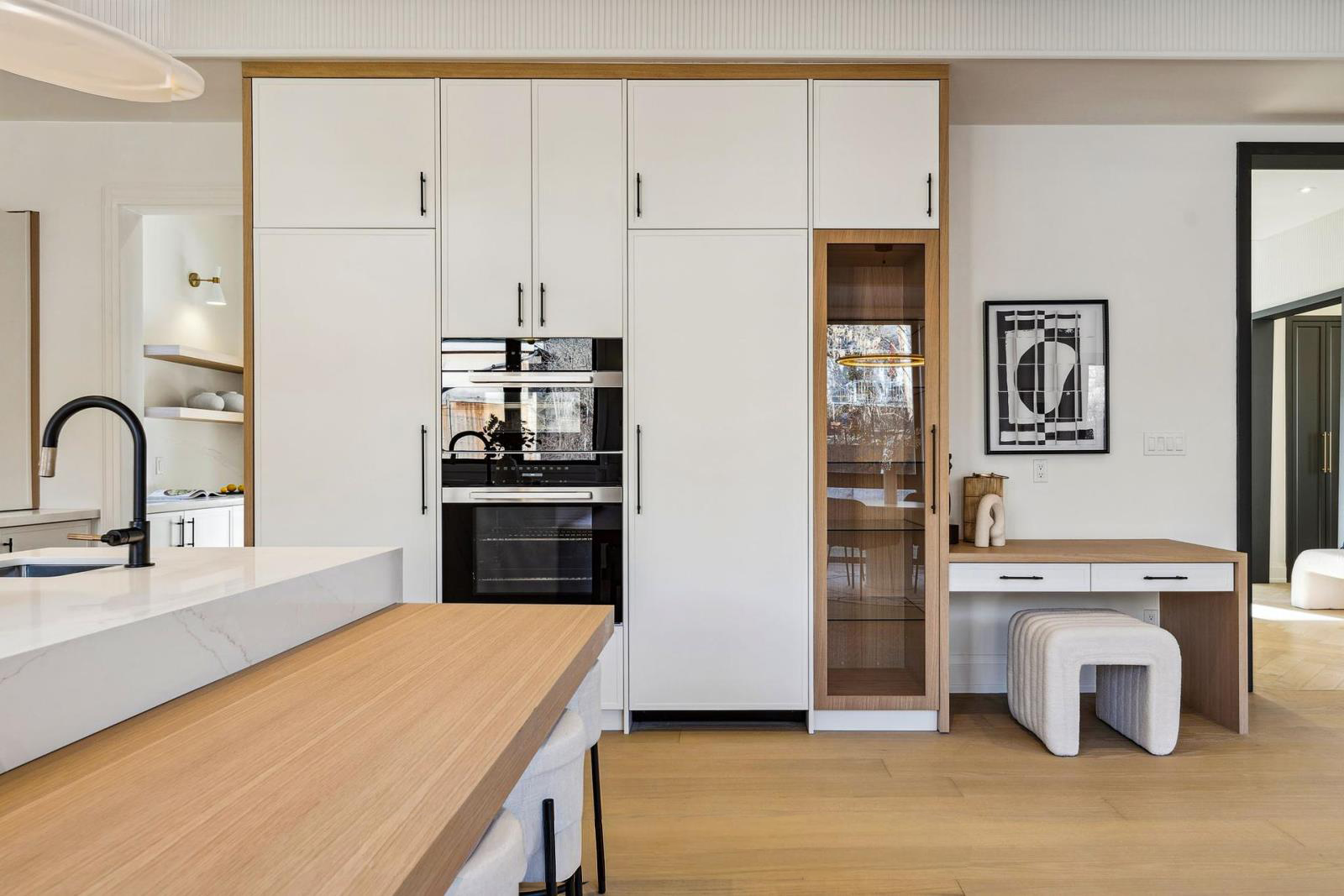 Modern kitchen with white cabinets, black handles, built-in oven, wooden accents, glass-front cabinet, abstract wall art, and a small desk with a white cushioned stool.