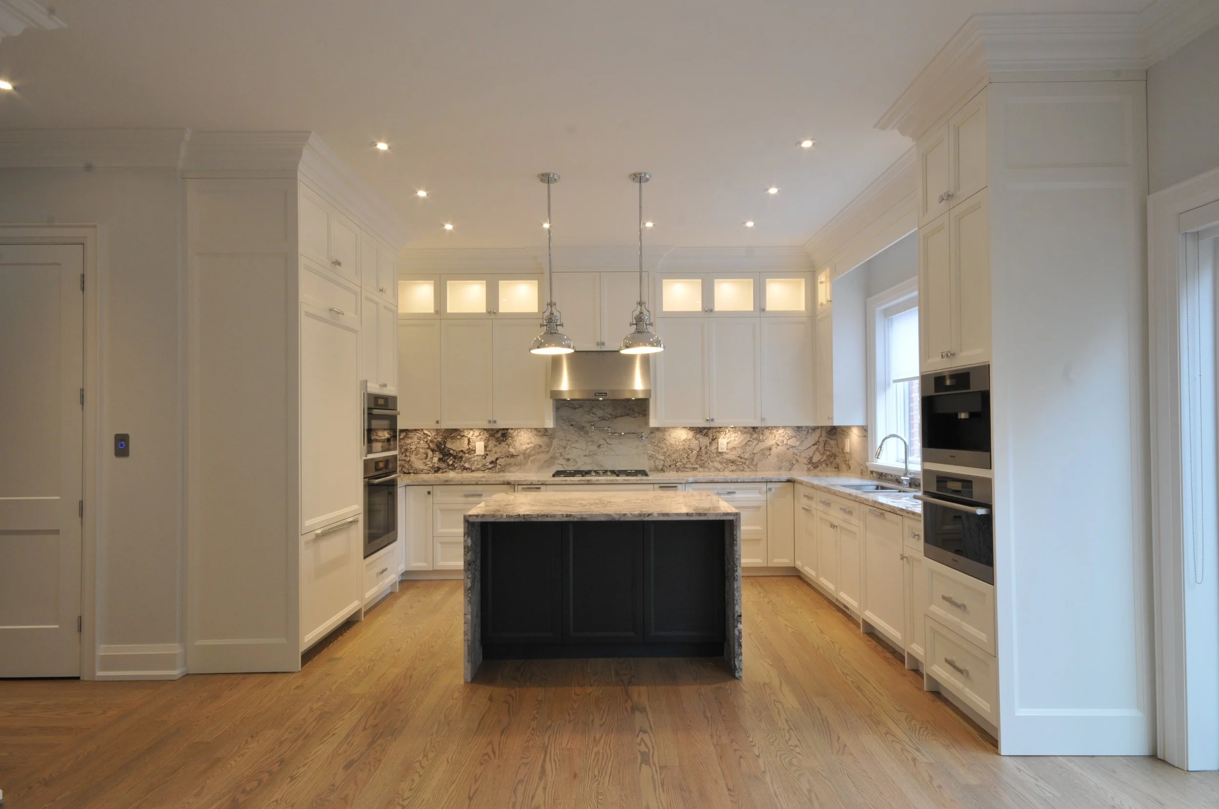 A modern kitchen with white cabinetry, a marble backsplash, and a black kitchen island with a marble top. Recessed lighting and two pendant lights hang over the island, and stainless steel appliances are integrated into the cabinets. Wood flooring co